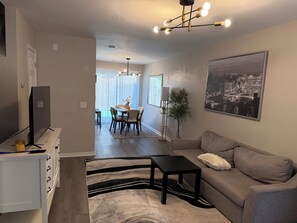 Una sala de estar moderna con un sofá, una mesa de centro y un televisor sobre un mueble. Se ve un área de comedor con una mesa y sillas al fondo.