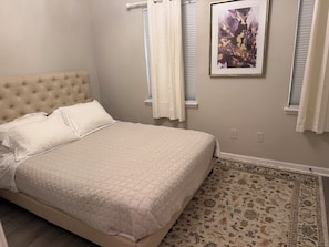 Une chambre à coucher avec un lit, une tête de lit capitonnée, un tapis à motifs et un tableau encadré au mur.