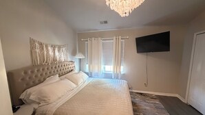 A bedroom with a tufted headboard, a large bed with white bedding, a wall-mounted TV, and a window with curtains. - Charming 2-bedroom townhome with WiFi and AC in Charlotte University Area (Charlotte)