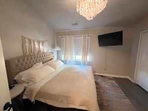 Un dormitorio con cabecera capitonada, una cama grande con ropa de cama blanca, un televisor montado en la pared y una ventana con cortinas.