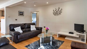 A modern living room with a leather sofa, a glass coffee table, a flat-screen TV, and a decorative wall piece. - Jubilee Barn Cottage at Bodmin Moor (St Breward)