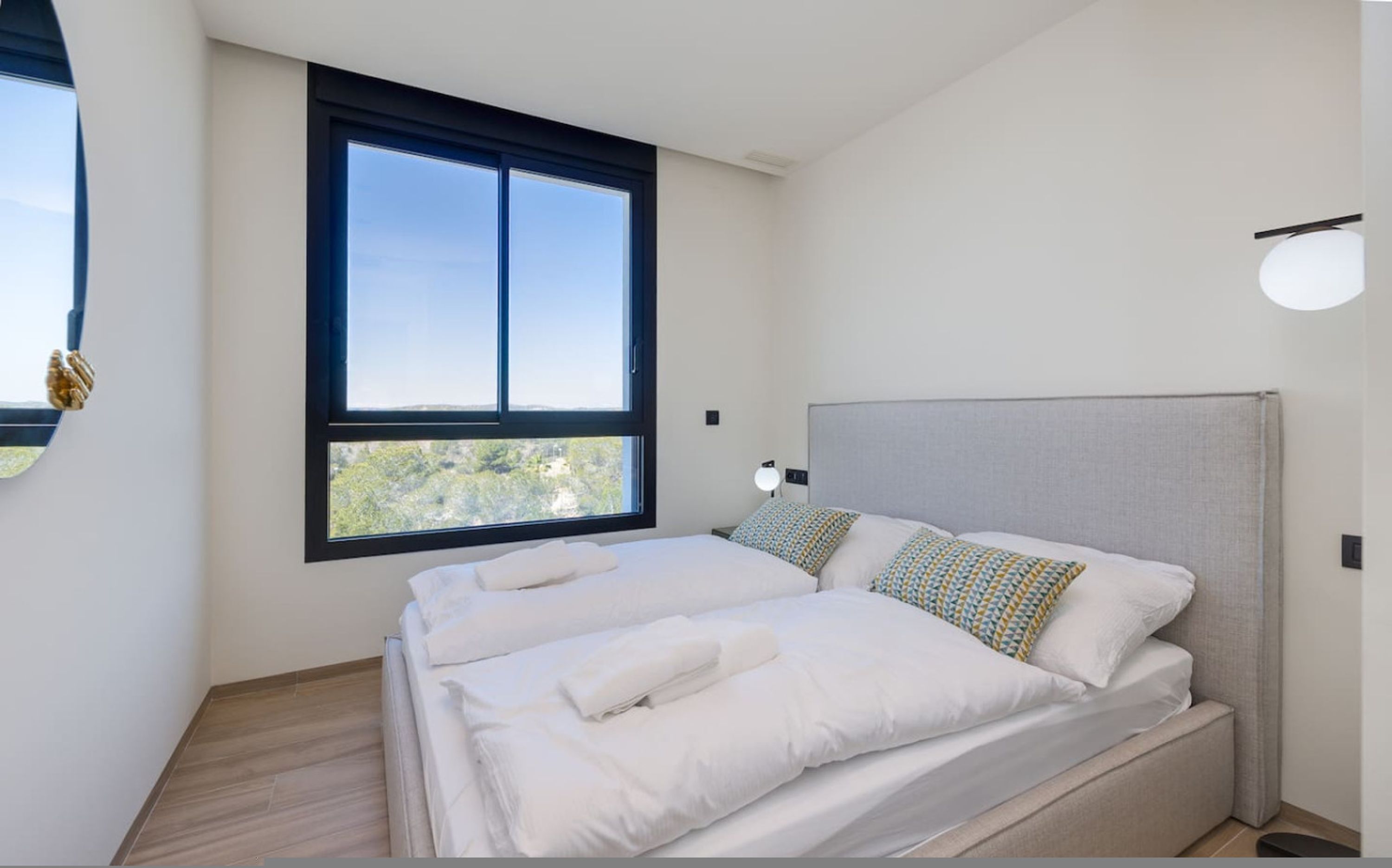 A modern bedroom with a large window, a bed with white linens, and a grey headboard.