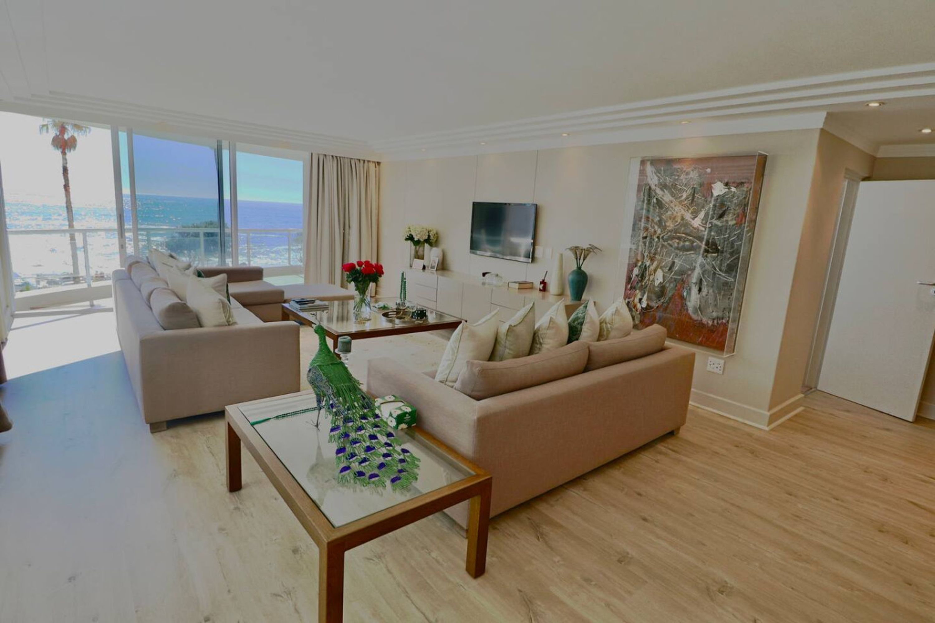 A modern living room with a sofa, a coffee table, and a TV. There is a large window with a view of the sea.