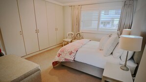 A bedroom with a bed, a bedside table with a lamp, a chair, a wardrobe, and a window with curtains. - Bantry Place - Luxury Apartment (Cape Town)
