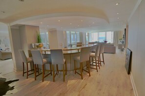 Un comedor moderno con una mesa redonda y seis sillas, una zona de cocina con un ventanal amplio, y una sala de estar con sofá y televisor.