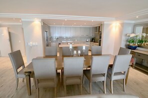 A modern dining area with a central table surrounded by chairs, a kitchen in the background, and a decorative vase with flowers on the right.