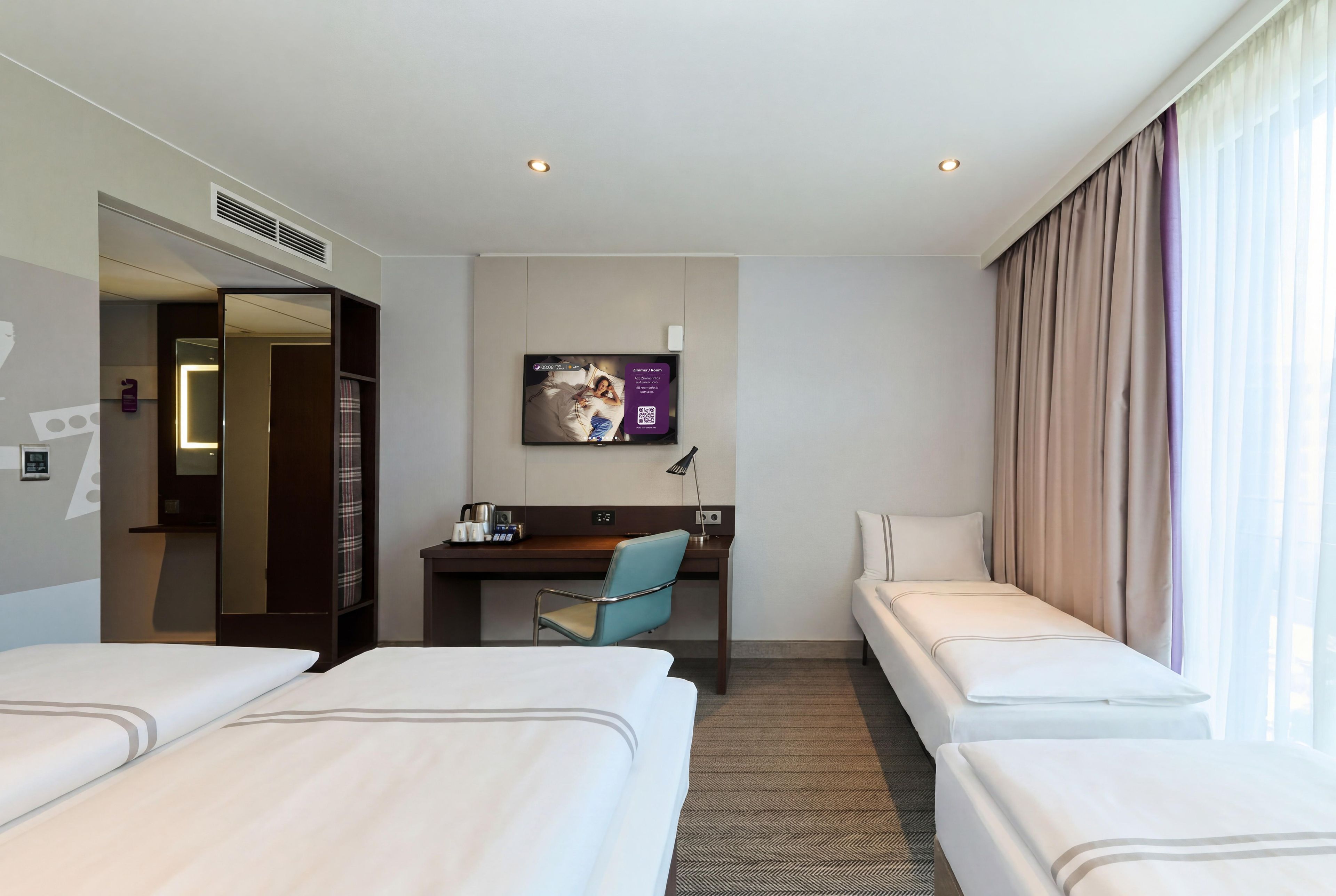 A hotel room with two single beds, a desk with a chair, a television, and a large window with curtains.