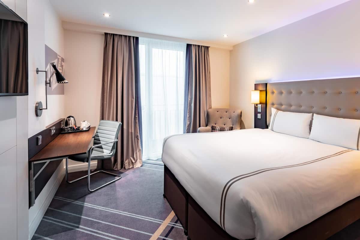 Habitación de hotel con una cama grande, un escritorio con silla, televisión y ventana con cortinas.