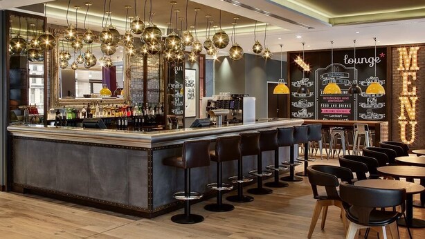 A bar area with a counter, stools, and hanging lights. - Premier Inn Frankfurt Messe (Frankfurt)