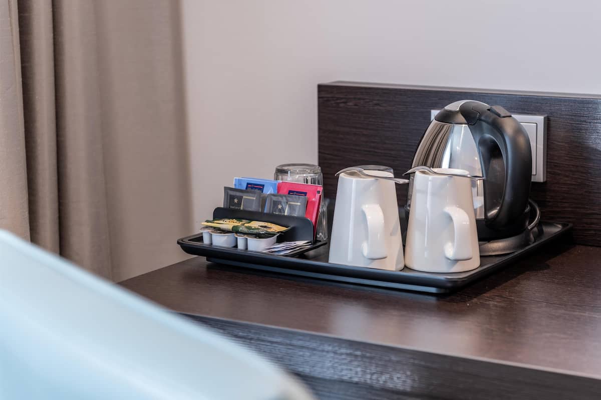 A hotel room with a dark wooden headboard, a tray with a kettle, two white mugs, and a set of toiletries.