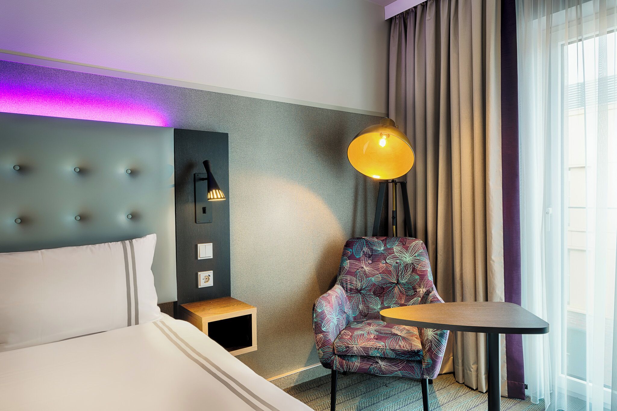 Habitación de hotel con cama, una silla, una mesa, una lámpara y un aplique de pared.