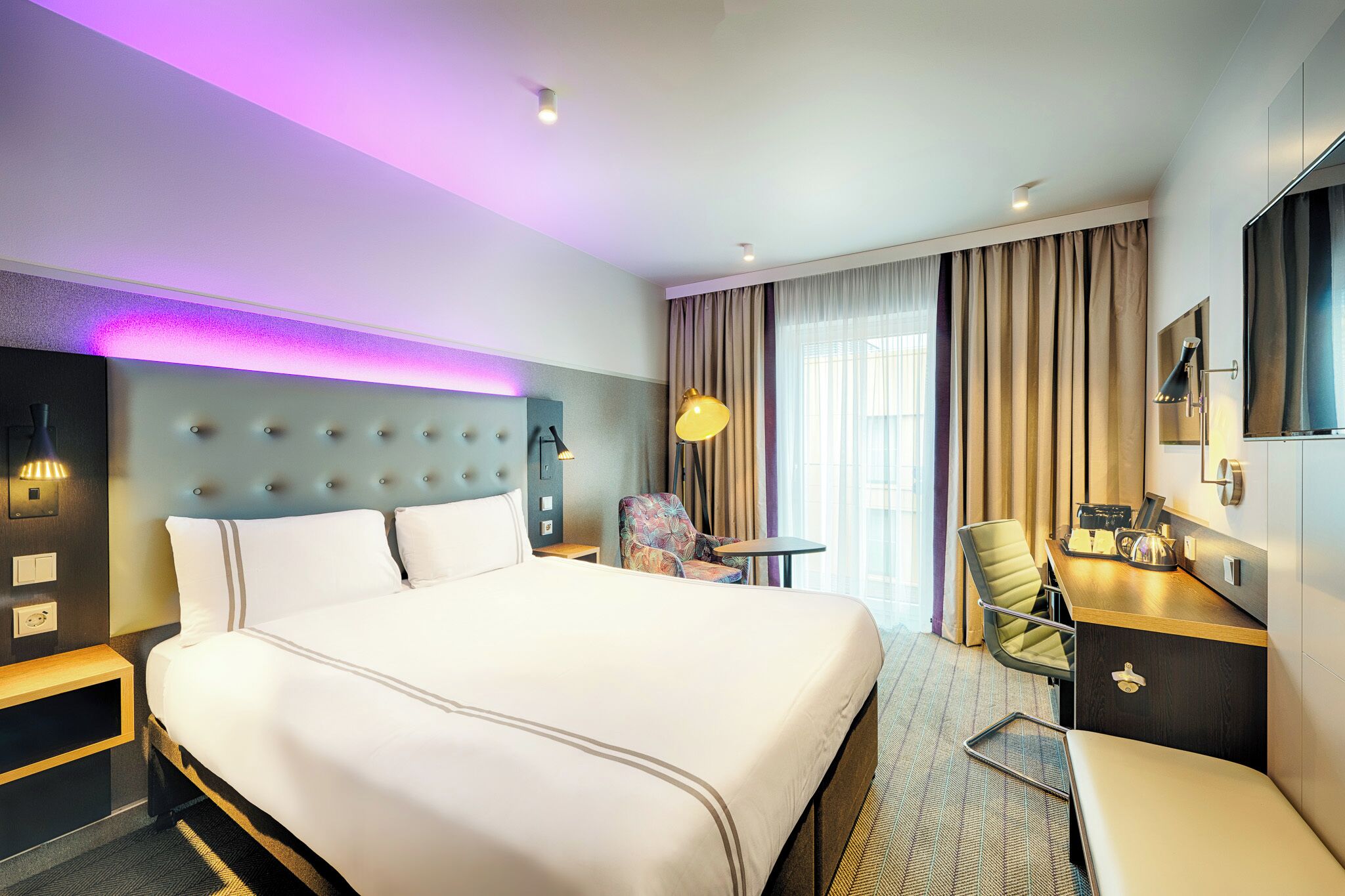 A hotel room with a large bed, a desk, a chair, a small table, and a window with curtains.