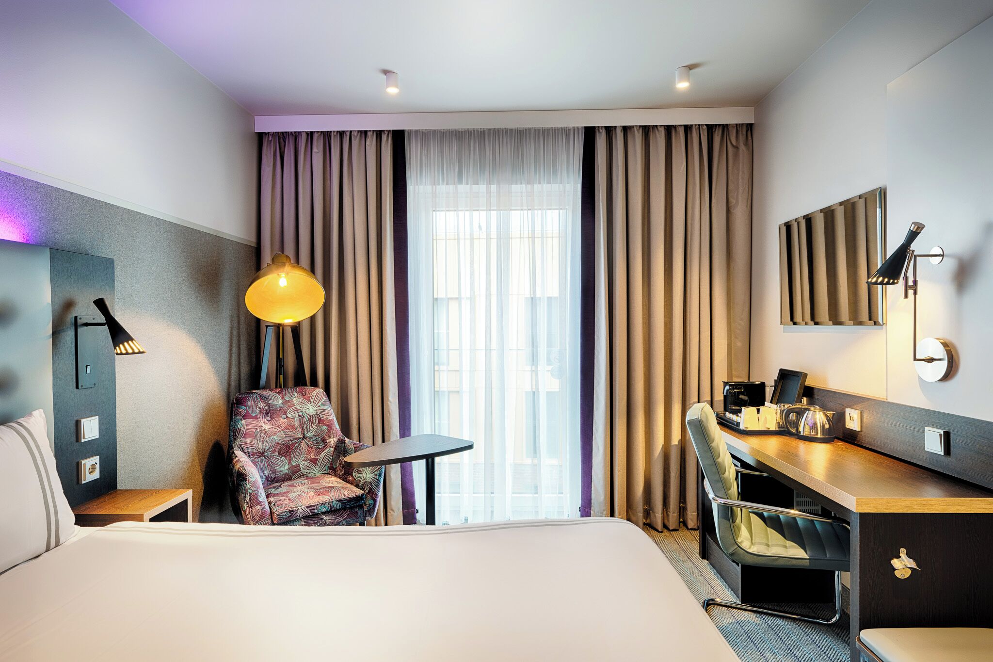 A hotel room with a bed, a desk, a chair, a lamp, and a view of the outside through large windows.