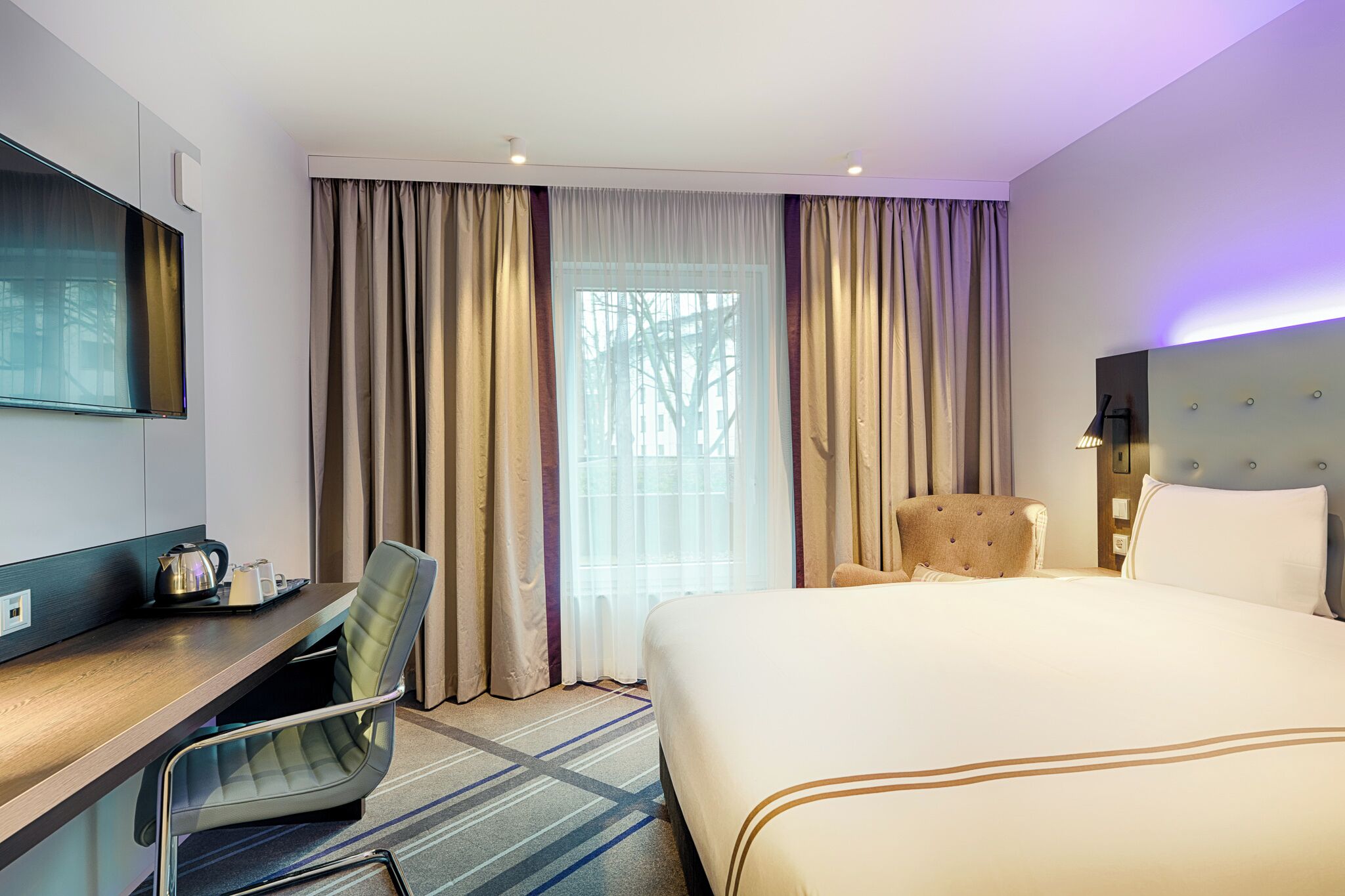 Habitació d'hotel amb llit, escriptori, cadira, televisió i finestra amb cortines.