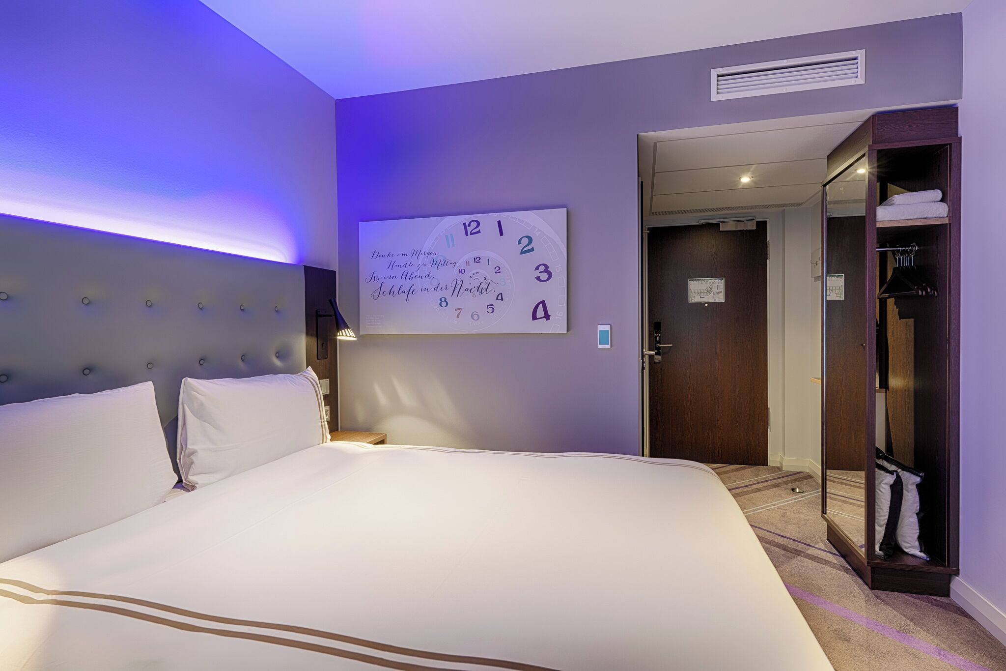 A hotel room with a large bed, a wall clock, a wardrobe, and a door leading to another room.