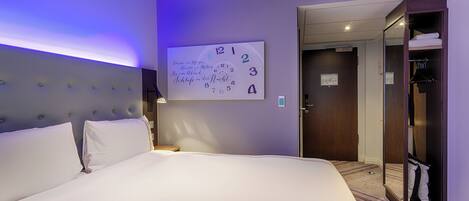 A hotel room with a large bed, a wall clock, a wardrobe, and a door leading to another room.