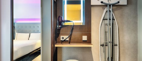 Habitación de hotel con cama, espejo con iluminación, secador de pelo, tabla de planchar y un taburete.