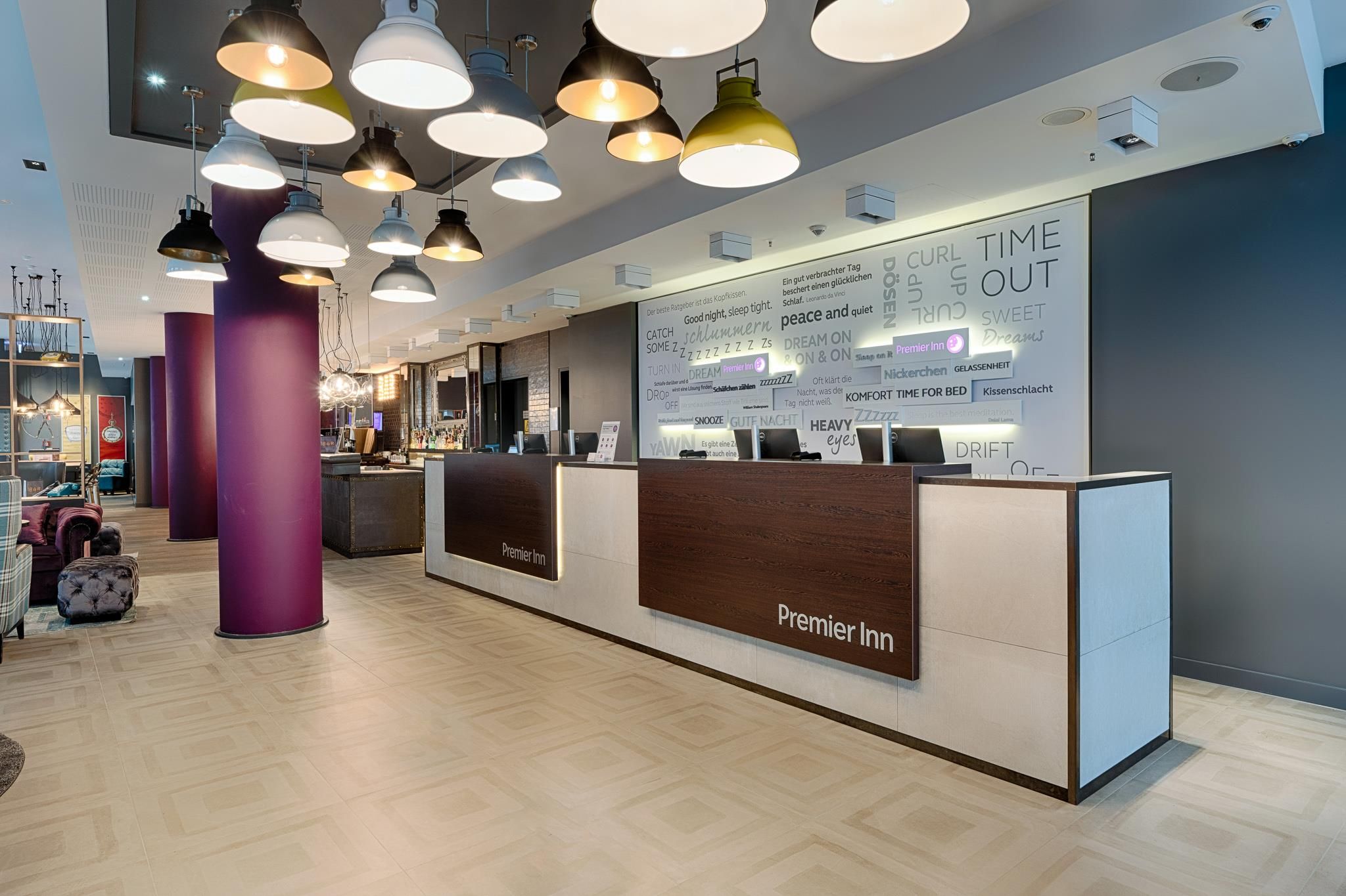 Zona de recepción de hotel con diseño moderno, que incluye un mostrador de recepción con el rótulo 'Premier Inn', una pared grande con texto e imágenes, y varias lámparas colgantes.