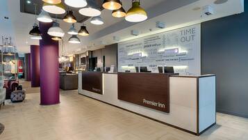 Zona de recepción de hotel con diseño moderno, que incluye un mostrador de recepción con el rótulo 'Premier Inn', una pared grande con texto e imágenes, y varias lámparas colgantes.