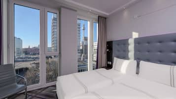 A hotel room with a large bed, a chair, and a view of a cityscape through floor-to-ceiling windows.