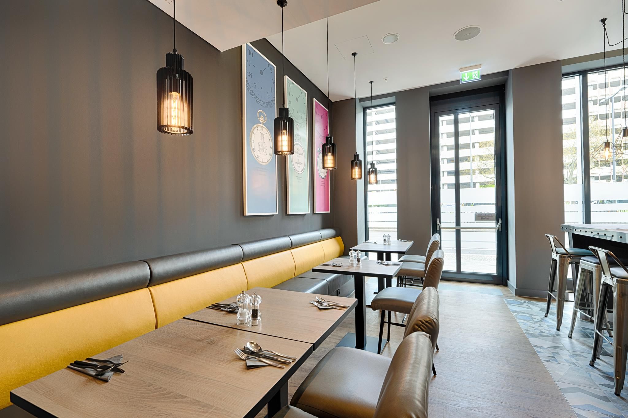 Een modern restaurantinterieur met een lange bankzitting, tafels met bestek, hanglampen en grote ramen.
