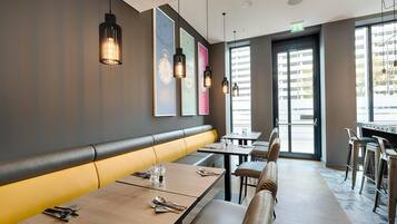 Een modern restaurantinterieur met een lange bankzitting, tafels met bestek, hanglampen en grote ramen.