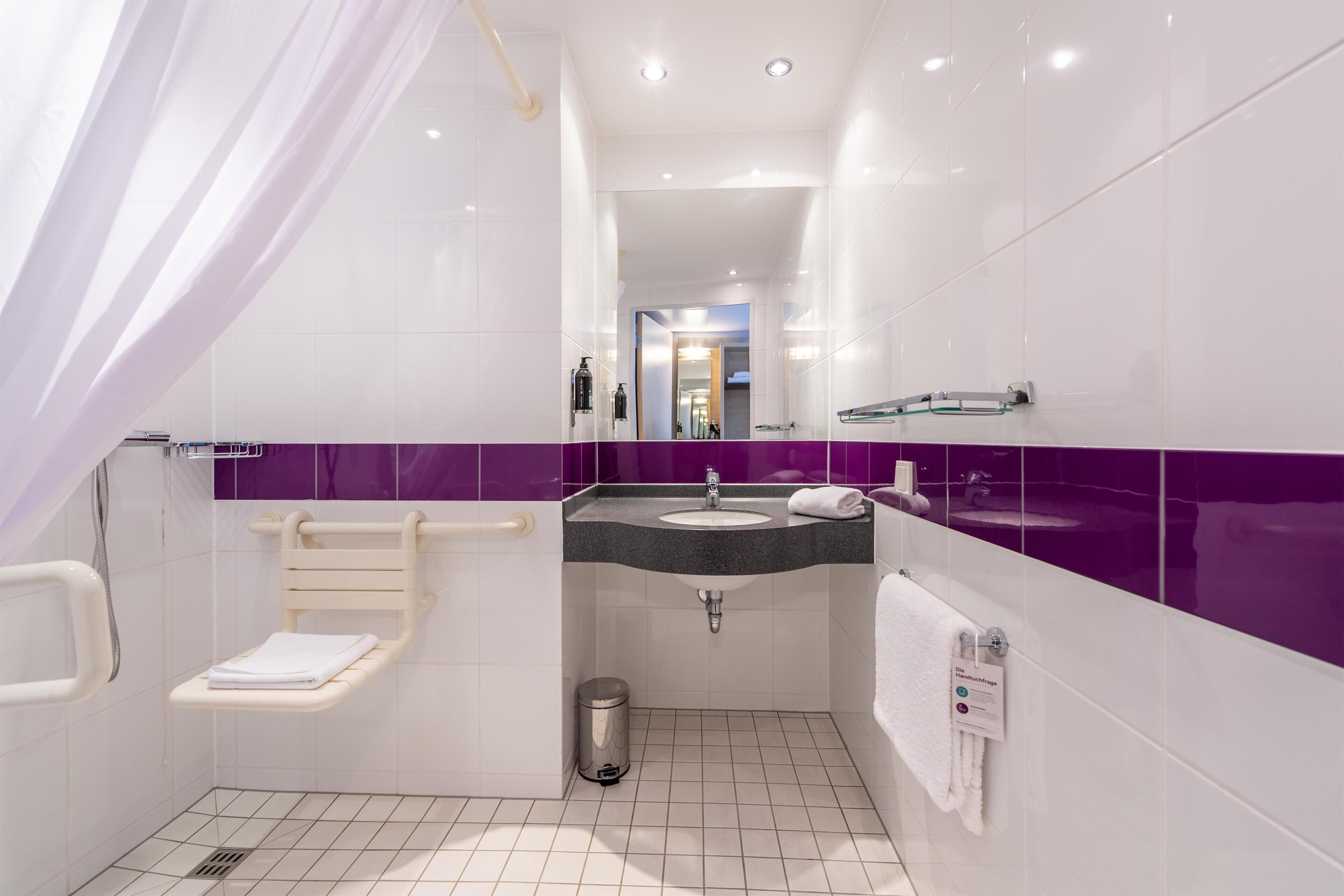 Baño moderno con ducha, inodoro y lavamanos, con azulejos blancos y morados, una barra de apoyo y un cubo de basura.