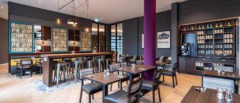 A modern restaurant with wooden tables and chairs, a bar area, and a bookshelf.