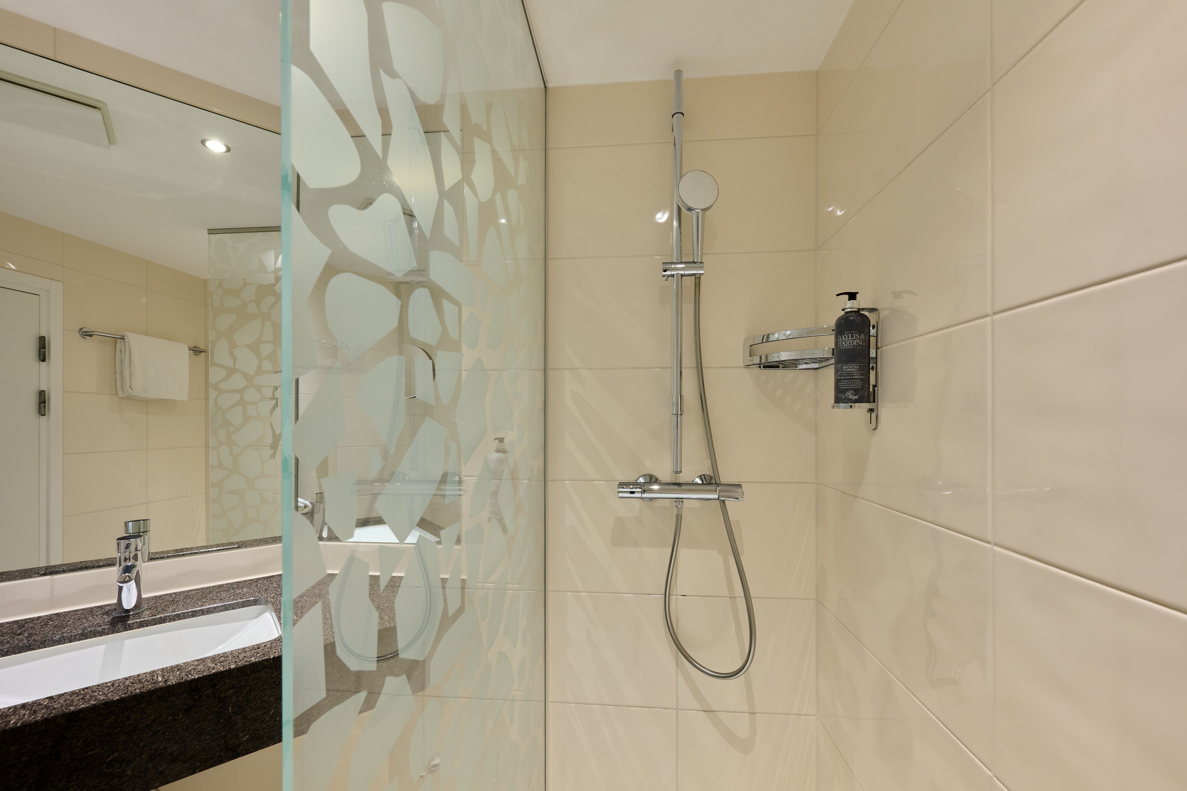 A modern bathroom with a glass shower enclosure, a handheld showerhead, a wall-mounted faucet, and a soap dispenser.