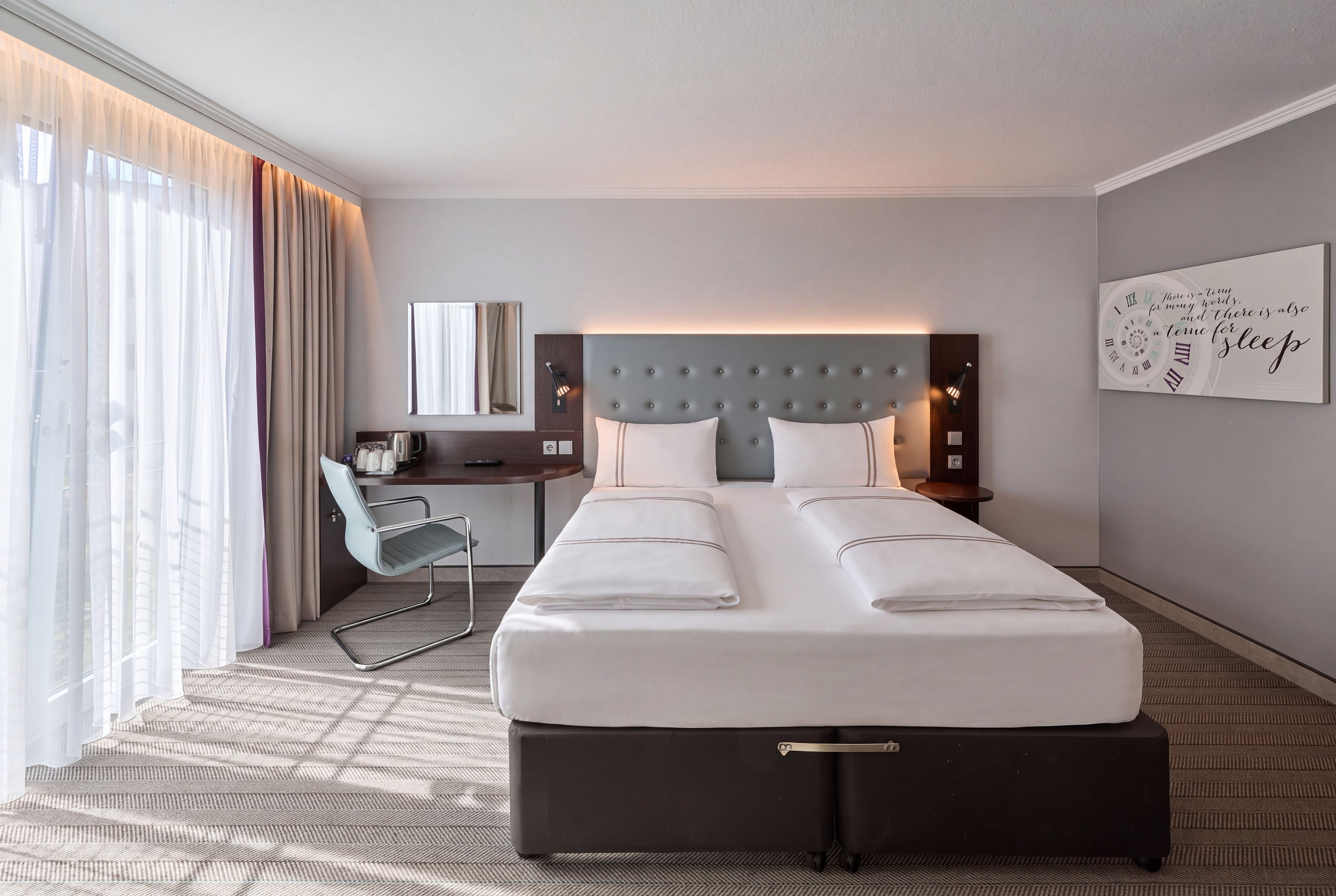 A hotel room with a bed, a desk, a chair, a bedside table, and a wall clock.