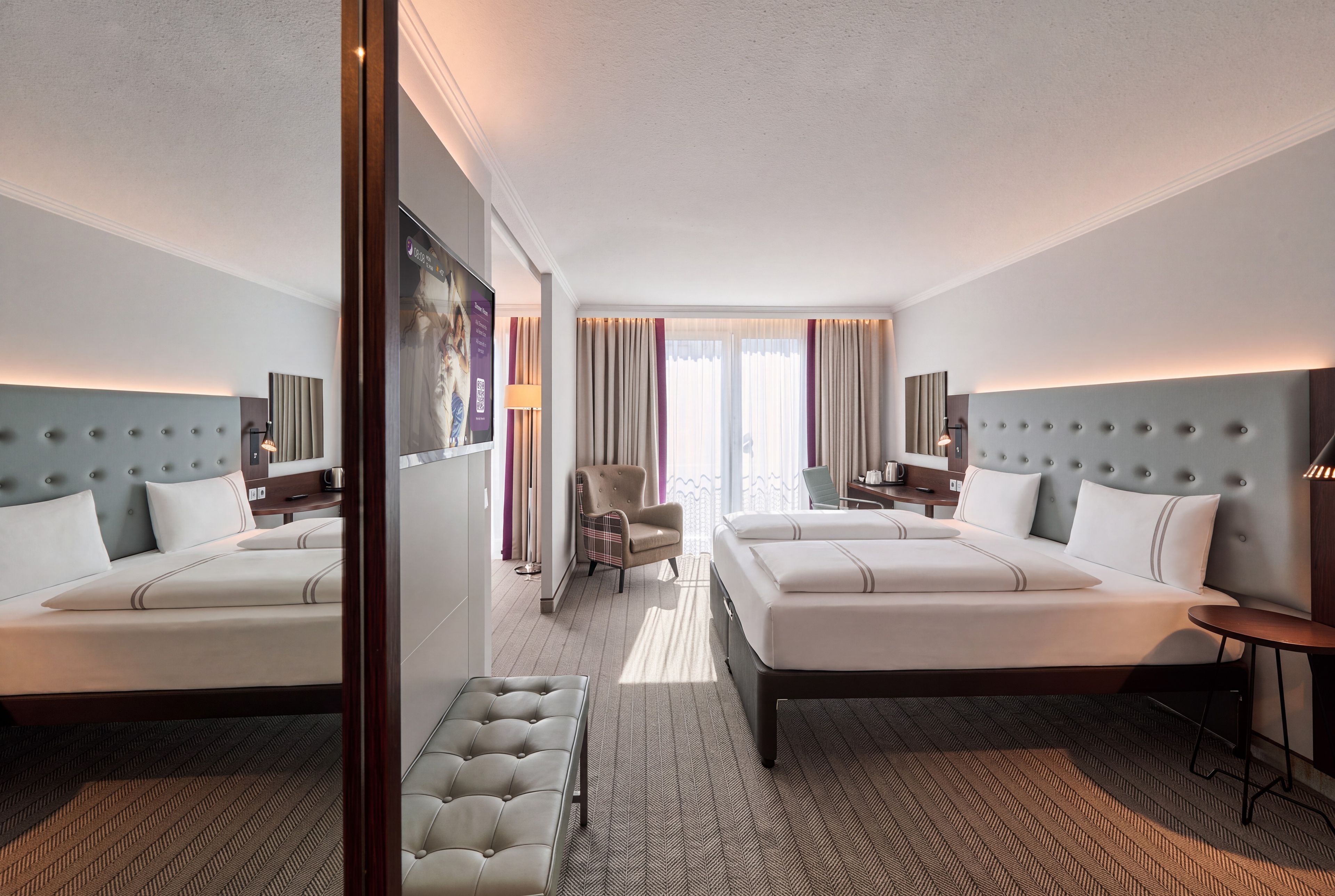 A hotel room with two beds, a sitting area, a desk, and a view of the outdoors.