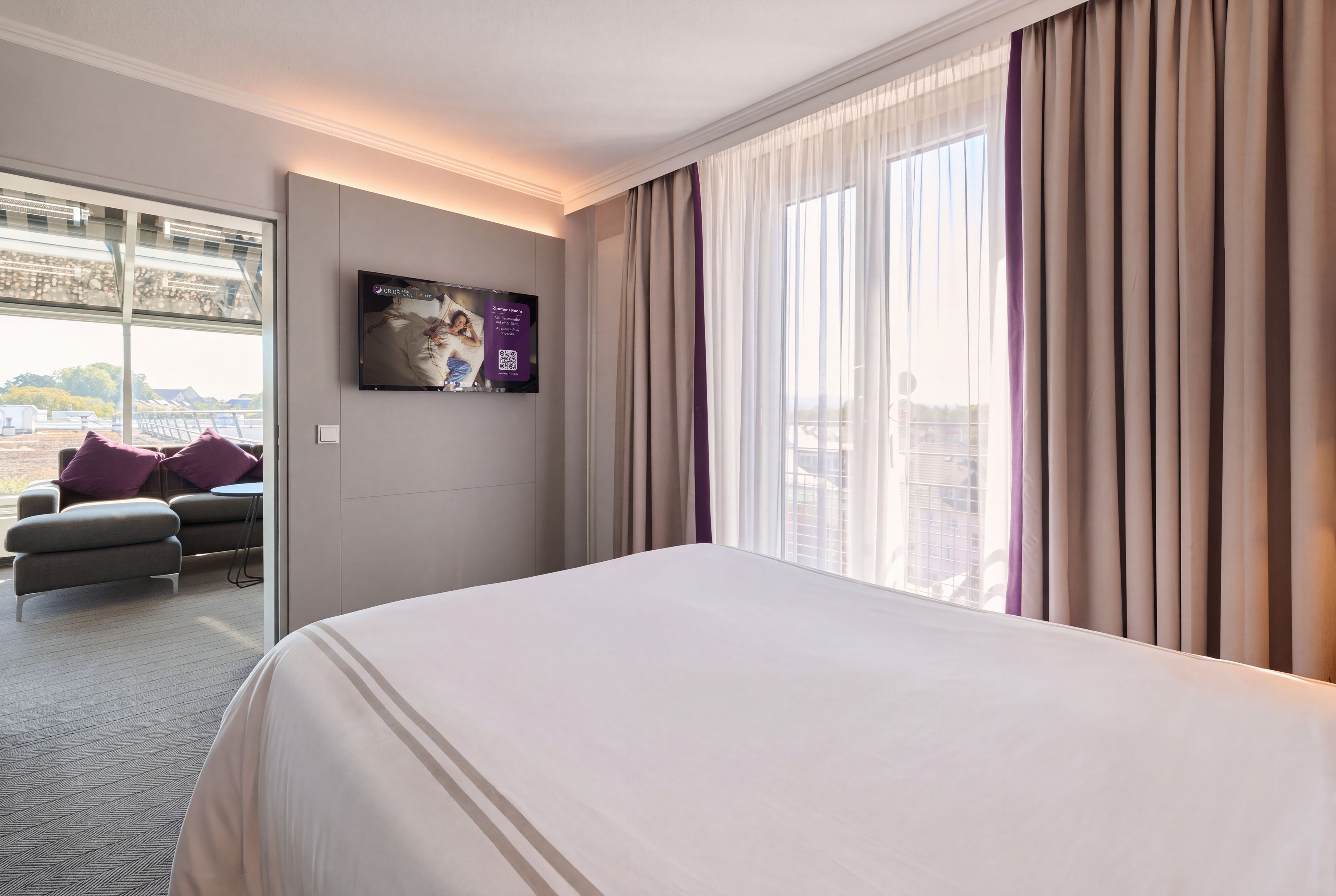 A hotel room with a bed, a television mounted on the wall, a window with curtains, and a view of the cityscape.