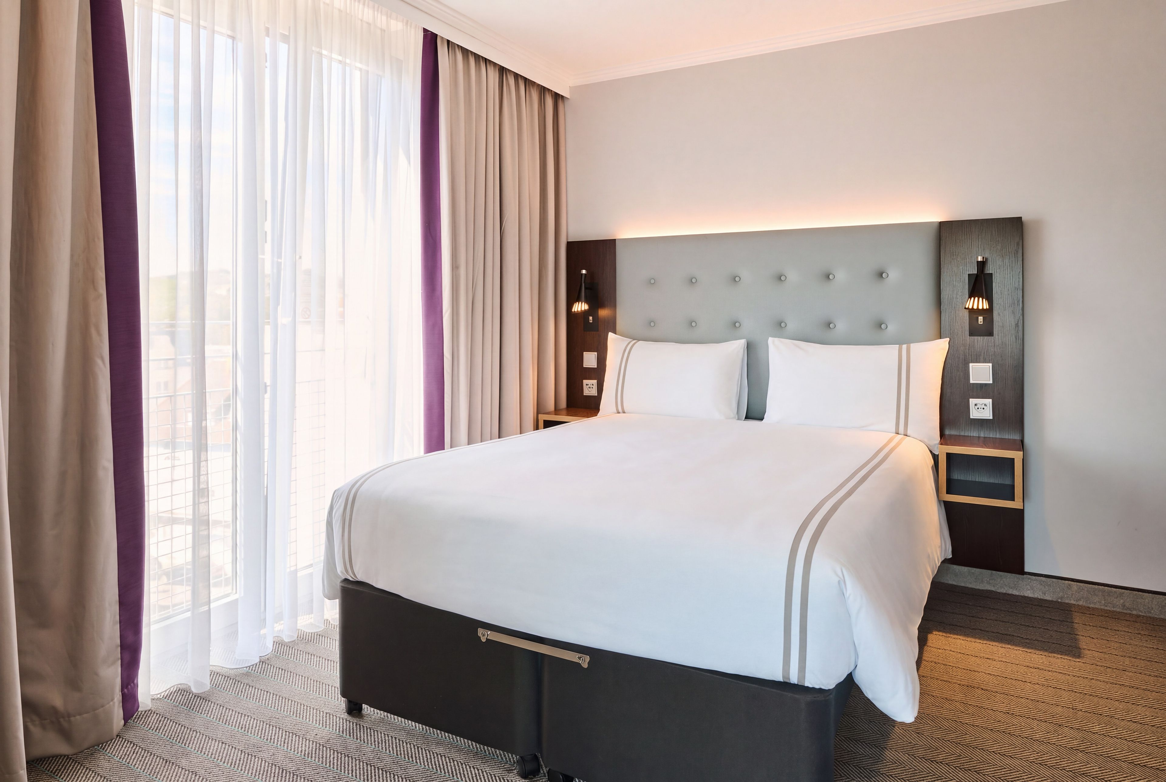 A hotel room with a large bed, a headboard with lights, a bedside table, and a view through the window.