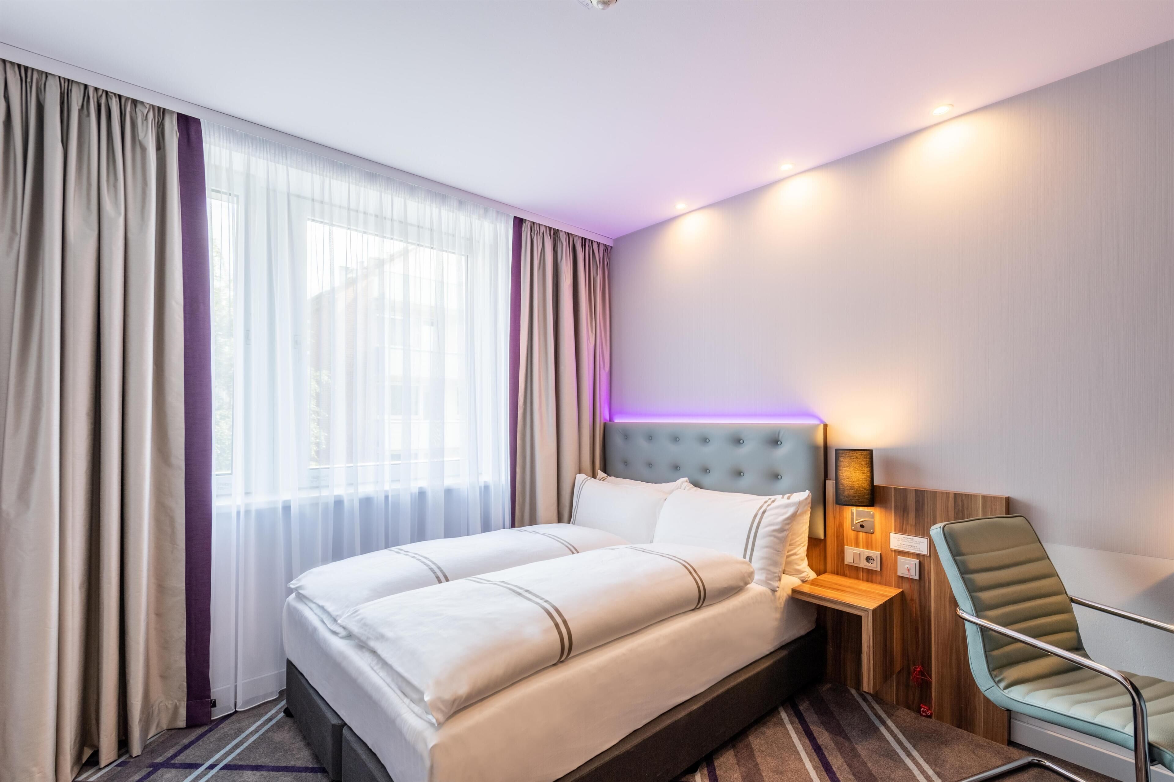 Habitación de hotel con una cama grande, una silla, una mesita de noche y un ventanal con cortinas.