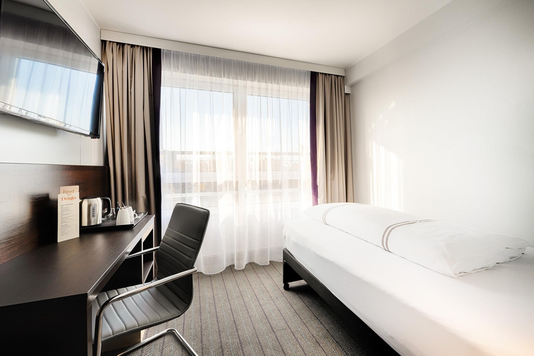A hotel room with a bed, a desk with a chair, a TV, and a window with curtains.