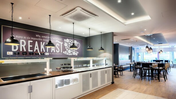 A modern breakfast bar with a large sign, pendant lighting, and a seating area with tables and chairs. - Premier Inn Berlin City Süd (Berlin)