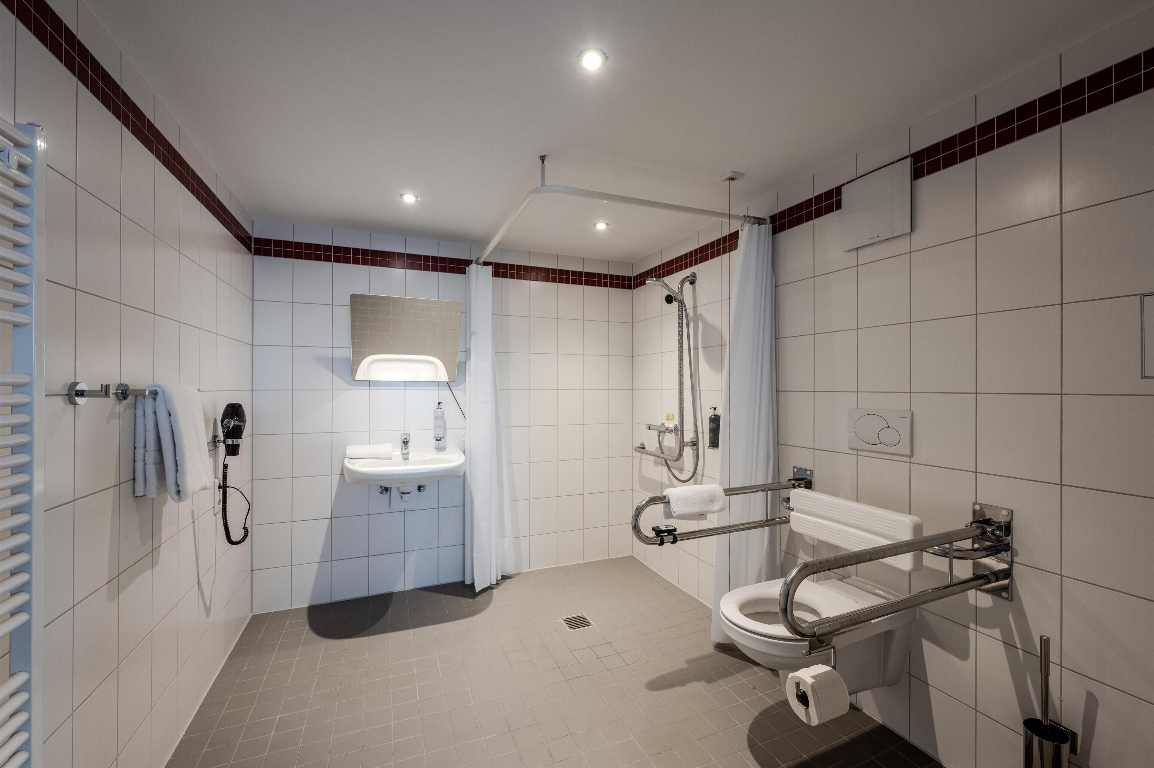 A modern bathroom with a shower, toilet, and sink, equipped with grab bars and a hand dryer.