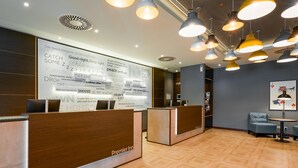 Hotellin vastaanottoalue, jossa on puusta valmistetut vastaanottotiskit, suuri seinä, jossa on tekstiä ja kuvia, sekä useita riippuvalaisimia.