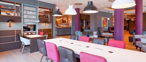 A modern hotel lobby with a long dining table, multiple seating areas, and a bar.
