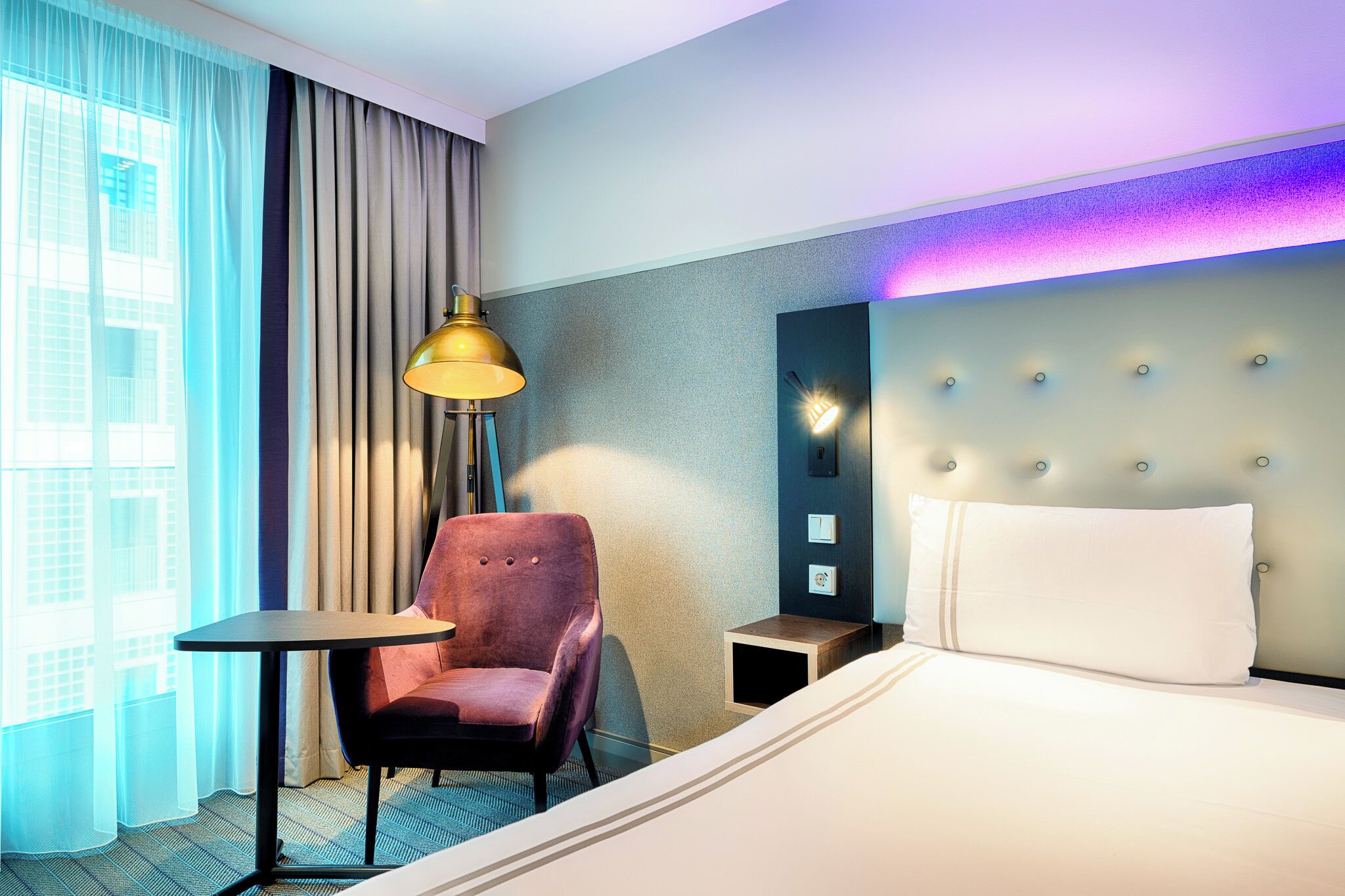 Une chambre d’hôtel comprenant un lit, une chaise, une table, une lampe, une table de chevet et un luminaire fixé au mur.