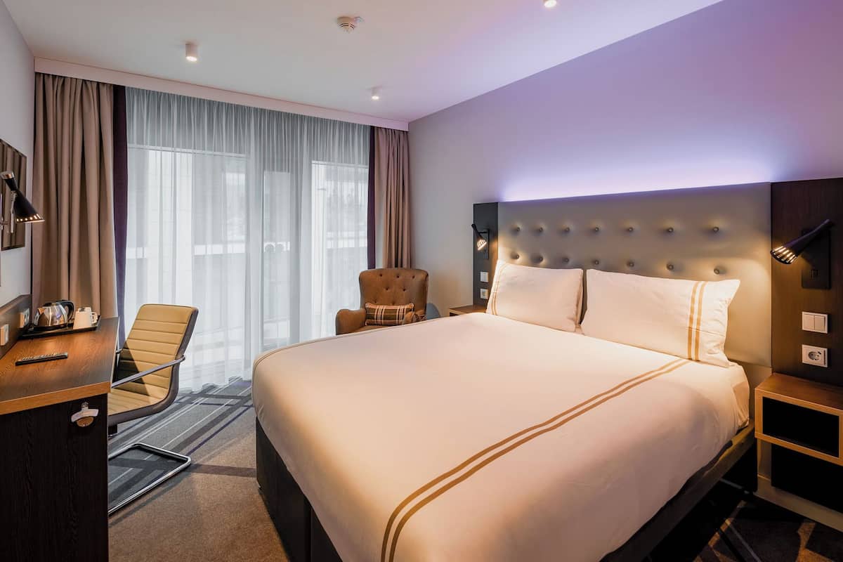 A hotel room with a large bed, a desk with a chair, a TV, and a wall-mounted light fixture.