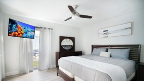 Ein Schlafzimmer mit einem Bett, einem Deckenventilator, einem Fernseher, einem Spiegel und einem Fenster mit Vorhängen.