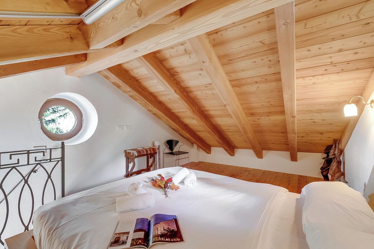 A bedroom with a wooden ceiling, a round window, a bed with white linens, a metal headboard, and a small table with a chair.