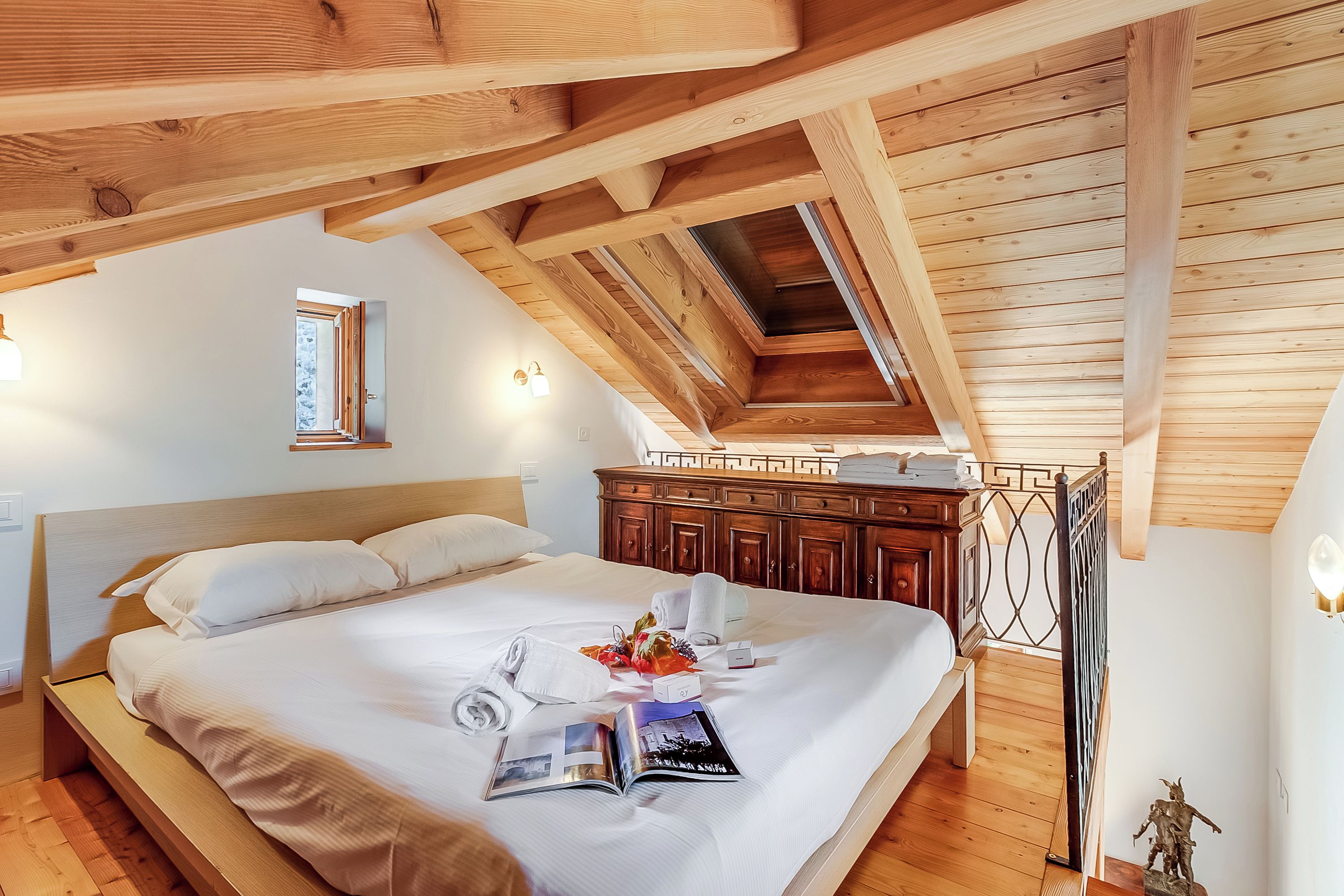 A neatly made bed with a magazine, towels, and a small statue on a wooden floor.