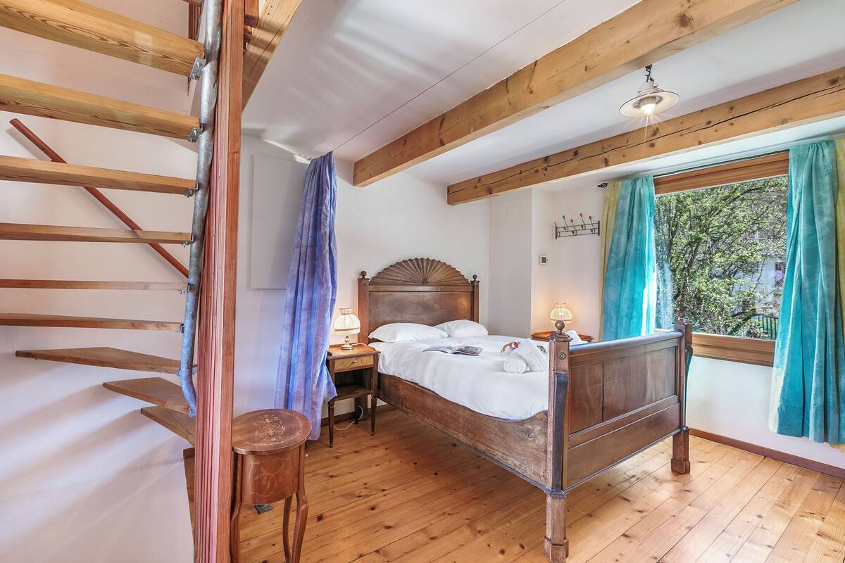 Ein schlicht eingerichtetes Schlafzimmer mit Holzbett, Nachttisch, Stuhl und einem Fenster mit Vorhängen.