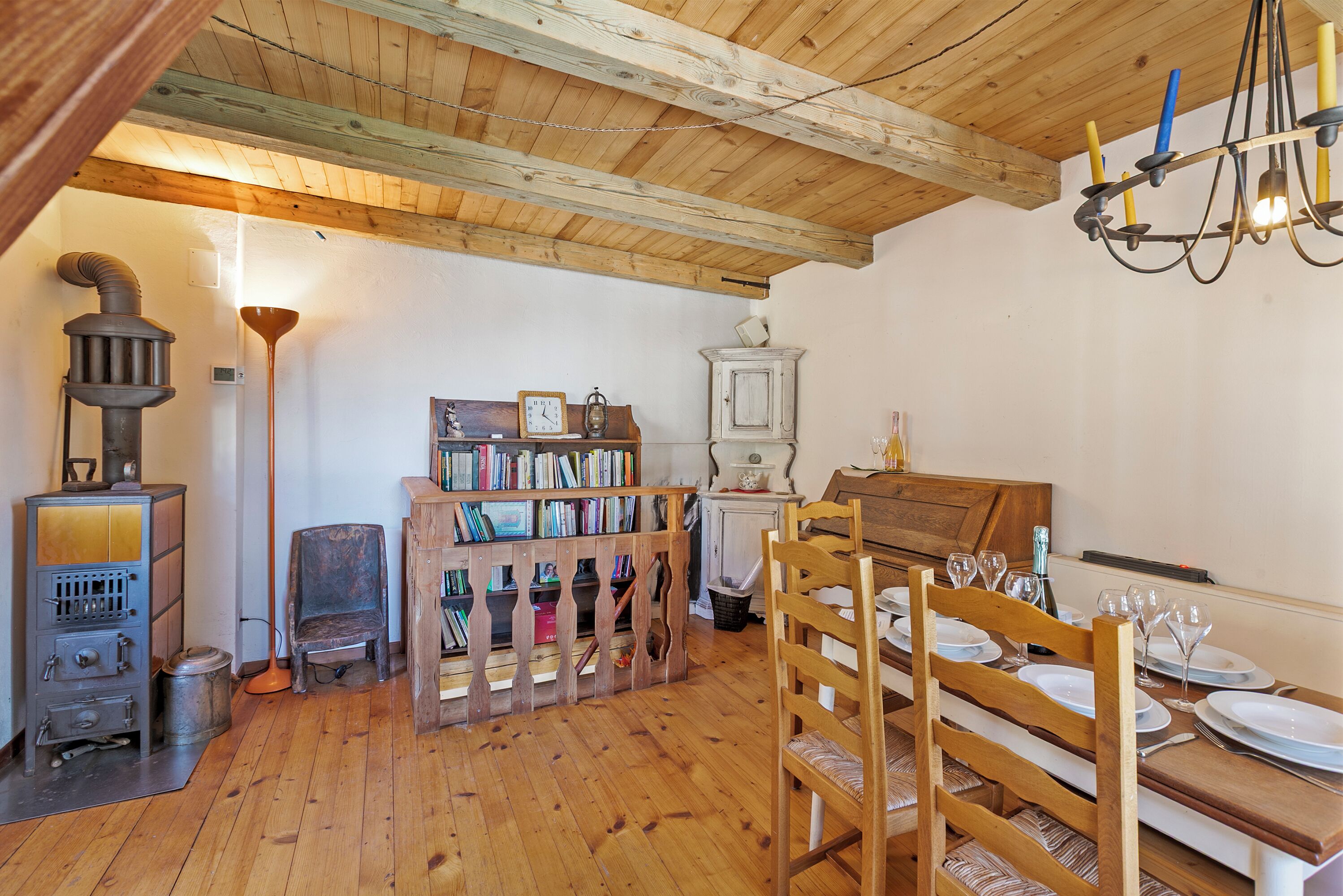 A wooden interior with a dining table set for four, a vintage stove, and a bookshelf filled with books.