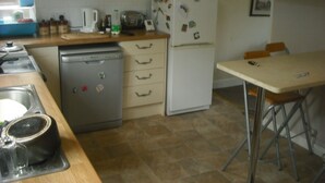 A compact kitchen with a dishwasher, microwave, refrigerator, and a small dining area with a table and chairs. - Room-large Home in Prime Location for al (Wetherby)
