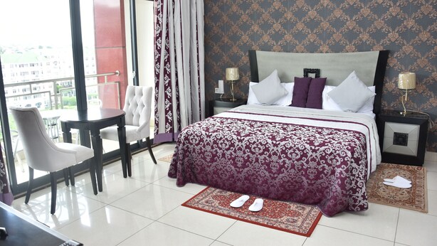 A hotel room with a large bed, two bedside tables, a chair, and a view of the city through large windows. - Hotel Carmen Galaxi (Malabo)