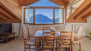 A dining area with a wooden table and chairs, a large window with a mountain view, and a ceiling with wooden beams. - Il Nido sul Lago by Varenna Holidays (Perledo)
