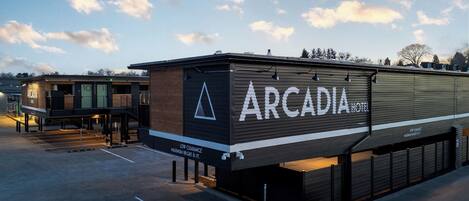 Un hotel moderno llamado "Arcadia Hotel" con un letrero llamativo, un área de estacionamiento techada y vistas al cielo.